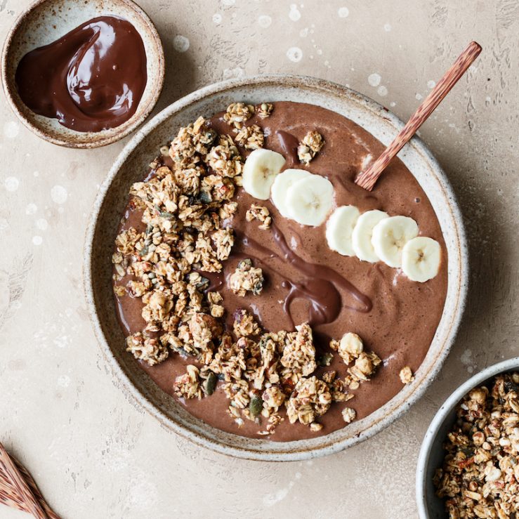 A bowl of chocolate and banana smoothie topped with slices of banana and granola and a drizzle of chocolate. A smaller bowl of melted chocolate and granola alongside the bigger bowl