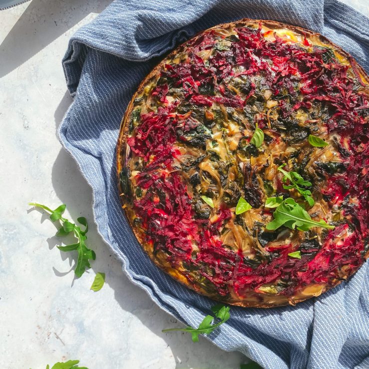 A moreish beetroot tart on top of a blue and white striped tea cloth with a couple of sprinkled rocket leaves