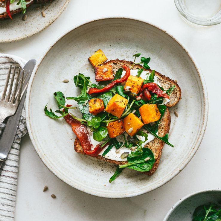 A grey plate with a slice of bread, roasted butternut squash and peppers and salad leaves with a knife and fork alongside