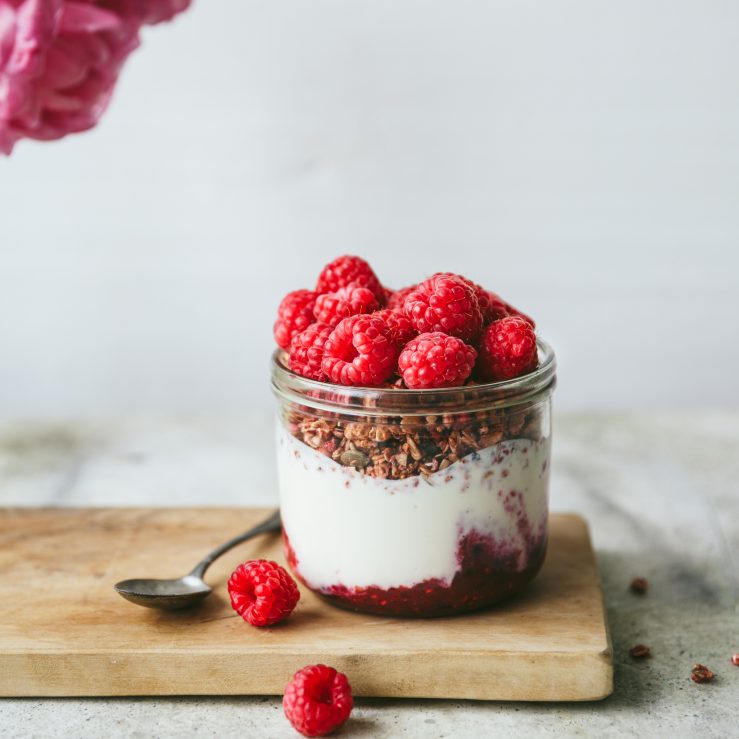 A glass pot of live yogurt with raspberry compot, granola and fresh raspberries on a wooden board with a spoon alongside
