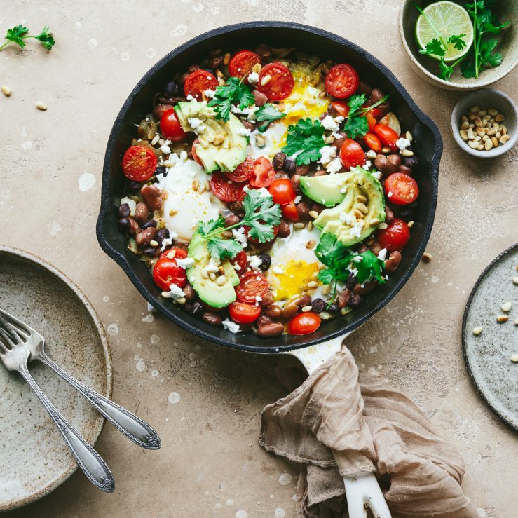 A saucepan showing a mixed bean shashuka topped with fresh tomatoes, yogurt, coriander, avocado and pine nuts