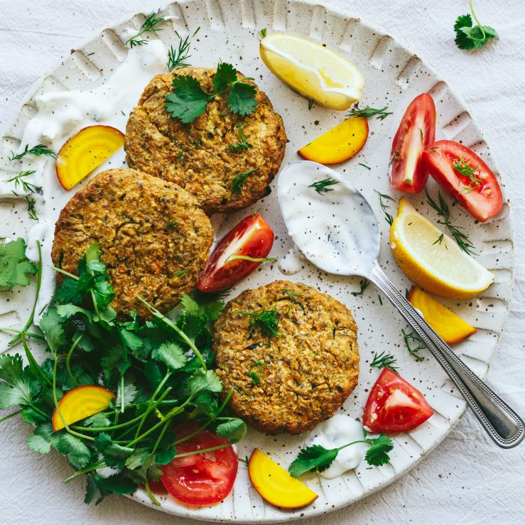 A large white platter with courgette and chickpea fritters, coriander sprigs, dill sprigs, tomatoes, lemon and probiotic tzatziki