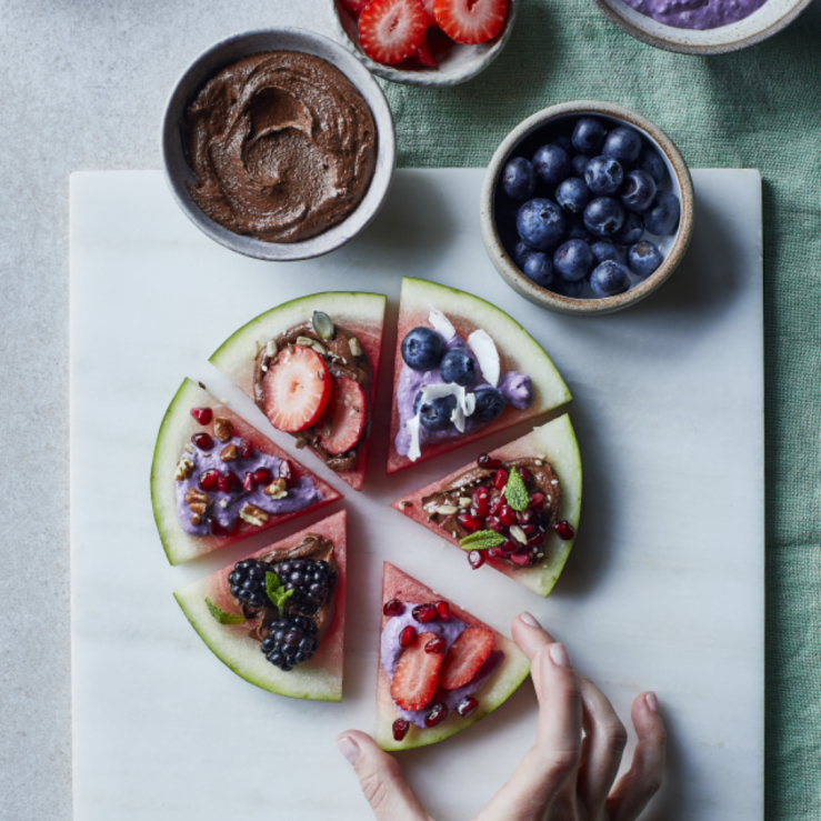 Loaded wedges of watermelon with blueberries, strawberries, yogurt and nut butter on a white slate