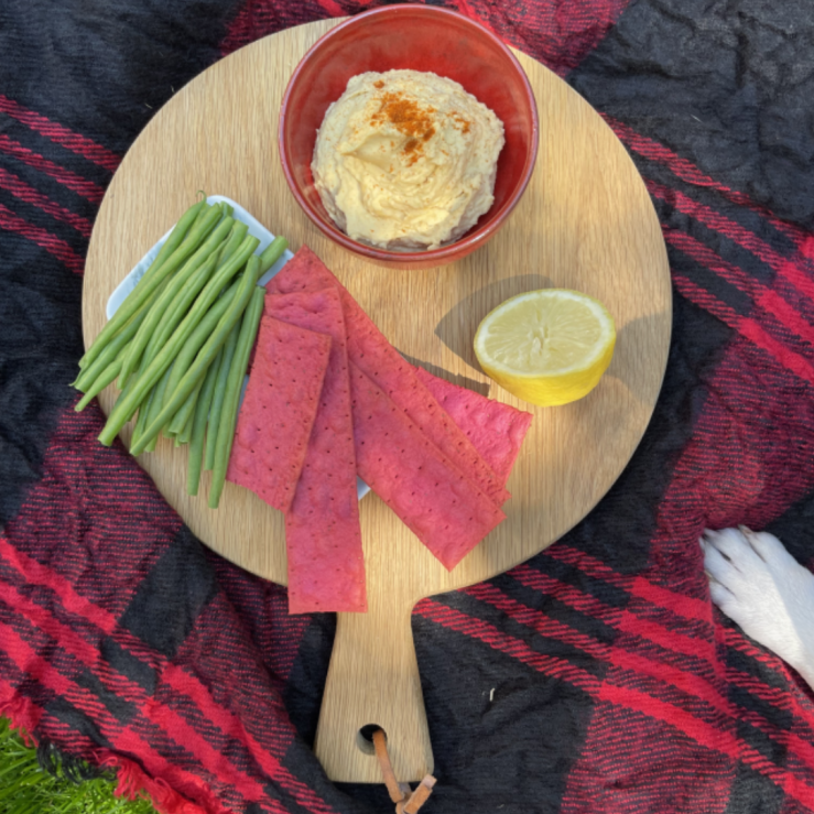 A wooden board on a picnic blanket with beetroot white bean hummus in a small bowl, half a lemon, beetroot crackers and fine green beans
