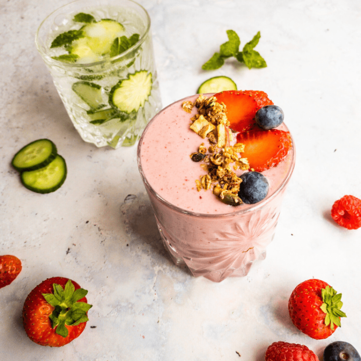 Two glass cups, one displaying berry smoothie with fresh fruit & the other with an elderflower cocktail mix with cucumber