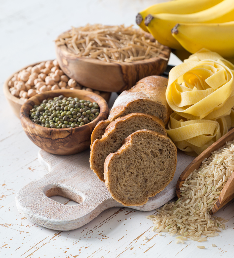 A white wooden board with bread, bananas, pasta and also rice, lentils and chickpeas in small wooden bowls displayed