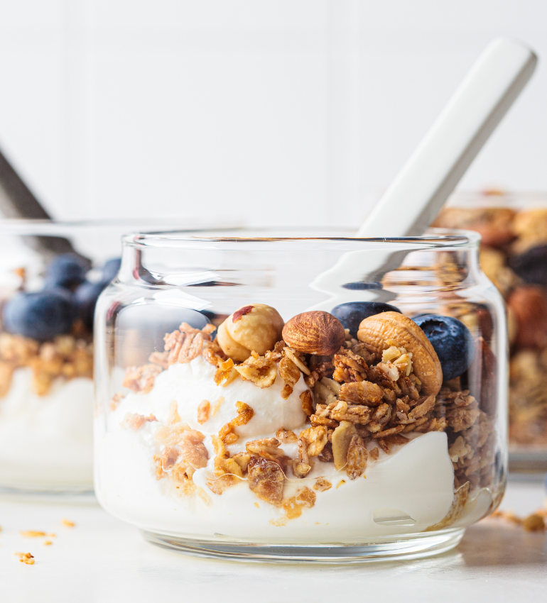 A glass bowl with a spoon filled with yogurt with granola, nuts and blueberries