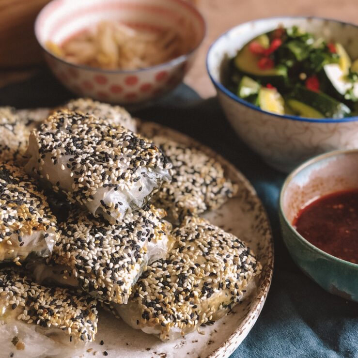 Rice paper dumplings covered in black and white sesame seeds on a plate with smaller plates in the background containing dips and salad