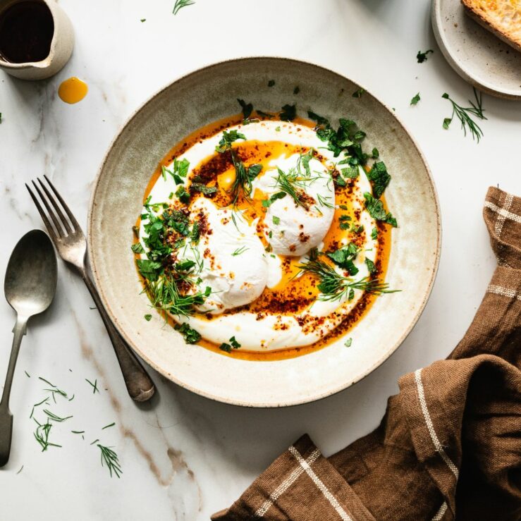 A bowl of prebiotic Turkish eggs topped with sprigs of dill and mint and a drizzle of chilli oil.