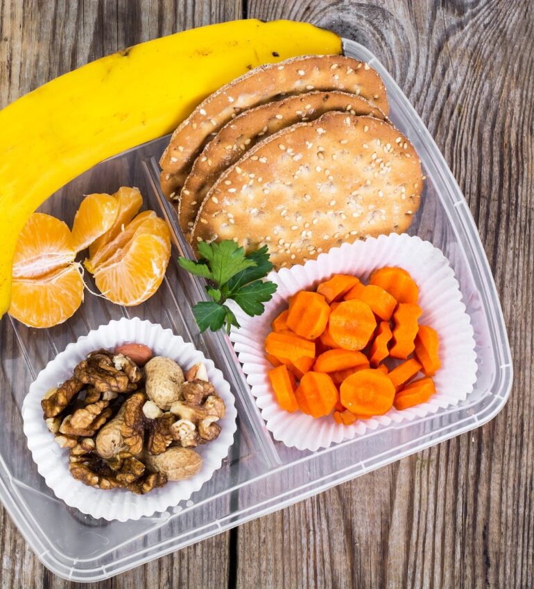 Tupperware box of snacks including a banana, crackers, carrots and nuts on a wooden bench for snacks to support gut health on holiday