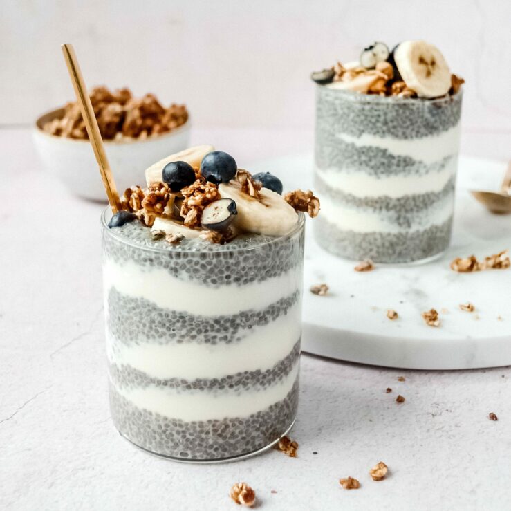 A picture of two clear glass tumblers with thick layers of plain yoghurt and chia seeds with a sprinkle of granola, sliced bananas and blueberries on top