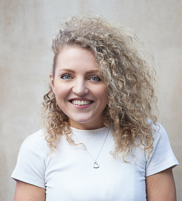 Image of a Lucy smiling, she has curly hair and wears a white t-shirt