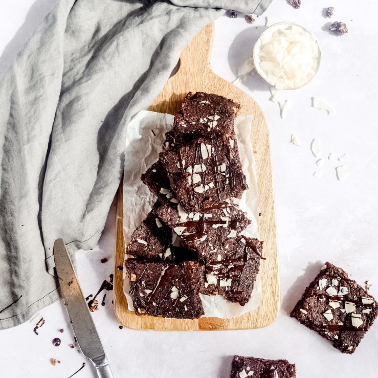 Eight chocolate and coconut brownies stacked along a chopping board, topped with coconut flakes