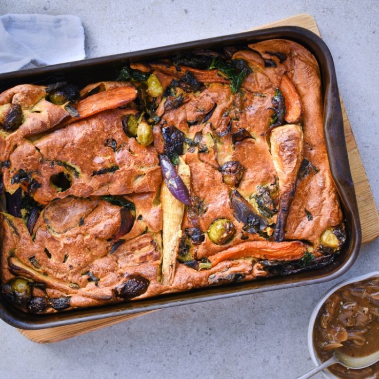 A baking tray on a table with the vegetable toad-in-the-hole recipe inside. It has lots of vegetables like carrots, parsnips and olives on it as well as a relish on the side.