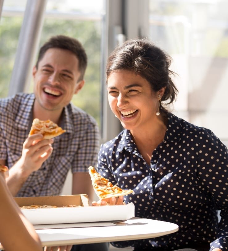 Two people sitting down, eating pizza and laughing