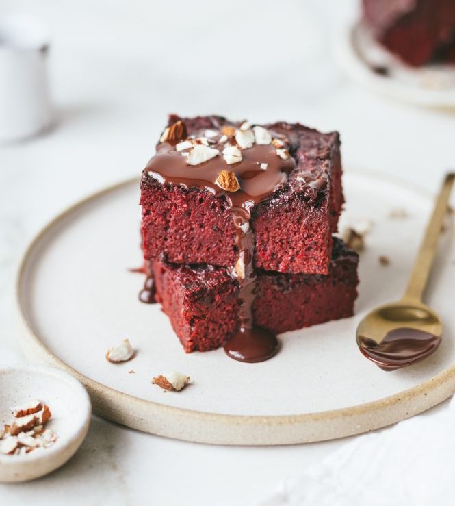 Two prebiotic beet velvet brownies stacked on a plate with melted chocolate and crushed almonds