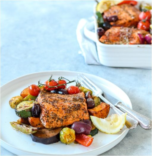 Dinner plate with Wild Alaska salmon fillet on top of traybake mixed roast vegetables, with larger tray of veg in background