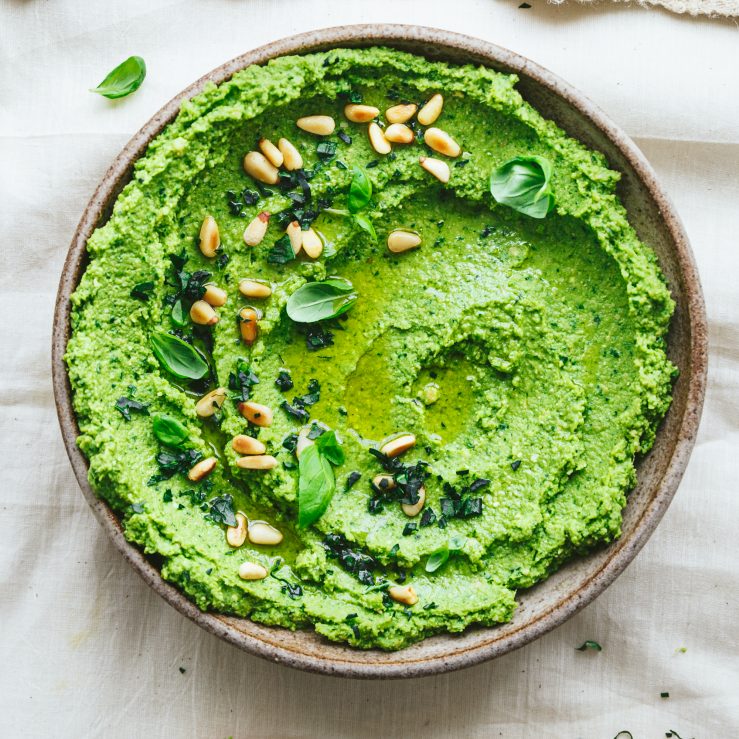 A bowl of leafy green pesto hummus with pine nuts and basil