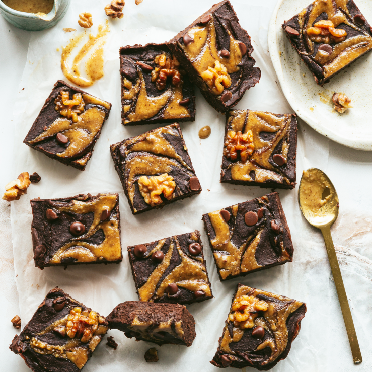 12 fudgy black bean and walnut brownies scattered on a bench top, drizzled with walnut butter