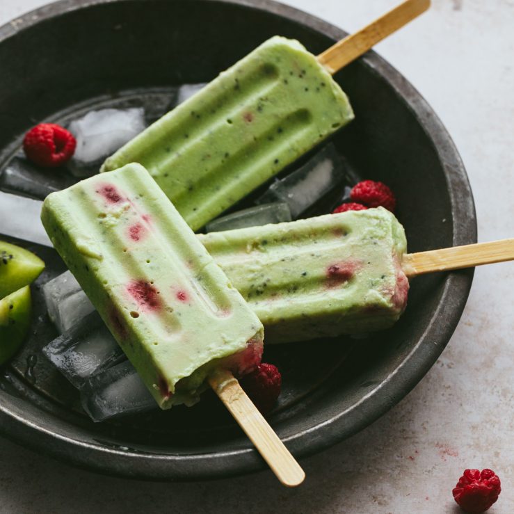 3 green ice lollies with raspberries in a black bowl with ice cubes